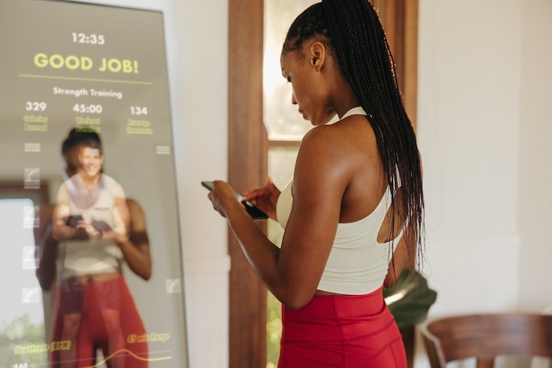 Using her smartphone to connect a smart mirror to a fitness app, a young female takes advantage of the latest in fitness technology to track her progress and stay motivated during her workout routine.