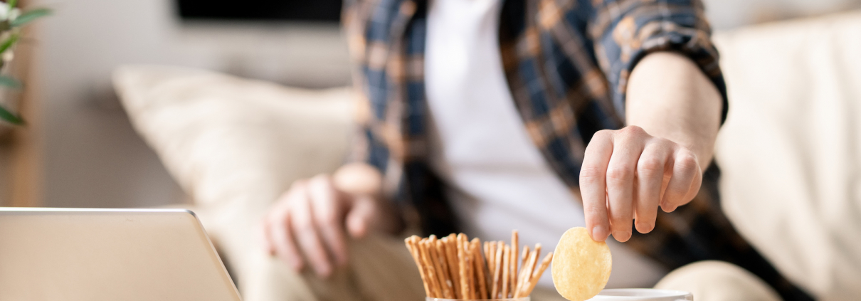 Young man taking potato chip out of glass bowl while sitting on sofa in front of laptop on table and having snack